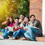 A group of college students sit together on campus steps smiling and studying with books and a laptop, representing the most prestigious summer programs.