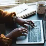 A student typing on a laptop with a notebook and coffee beside them, representing a participant drafting creative work for the Kenyon Young Writers Workshop.