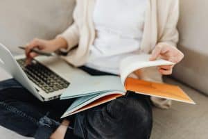 A person sitting on a couch typing on their laptop while flipping through a book