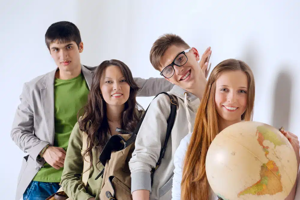 Group of cheerful happy students holding world globe and looking at camera leaning on white wall at campus.