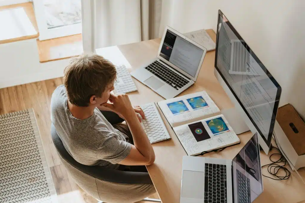 A man sitting on a desk surrounded by computers and learning materials