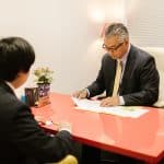 Harvard admissions consultant in a suit and gold tie reviewing documents with a student at a red desk