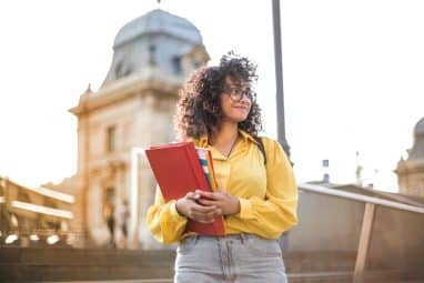 A young female student with curly hair and glasses holds a binder and smiles thoughtfully on a university campus, considering how to stand out to colleges.
