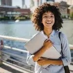 Smiling student holding a laptop outdoors with city buildings in the background, symbolizing preparation for the Harvey Mudd application deadline.