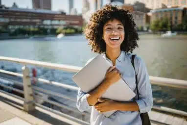 Smiling student holding a laptop outdoors with city buildings in the background, symbolizing preparation for the Harvey Mudd application deadline.
