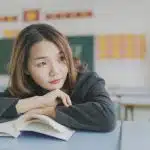 A student sits at her desk with an open book, thoughtfully considering colleges still accepting applications.