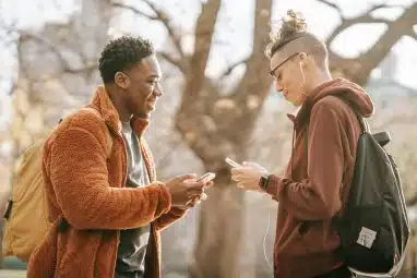 Two college students wearing backpacks smile and talk while looking at their phones outdoors, illustrating the excitement of preparing before the Wake Forest application deadline.