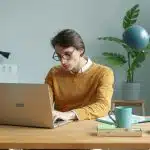 A student wearing glasses and a mustard sweater studies on a laptop at a wooden desk with plants and notebooks nearby, researching the Boston College application deadline.