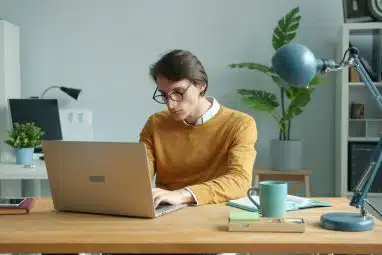 A student wearing glasses and a mustard sweater studies on a laptop at a wooden desk with plants and notebooks nearby, researching the Boston College application deadline.
