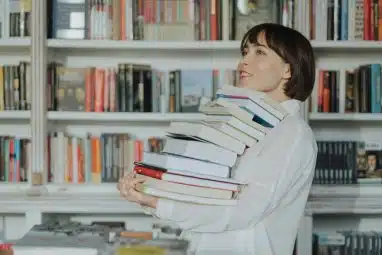 Woman carrying a tall stack of books in a library, illustrating study preparation for Georgetown SAT requirements.