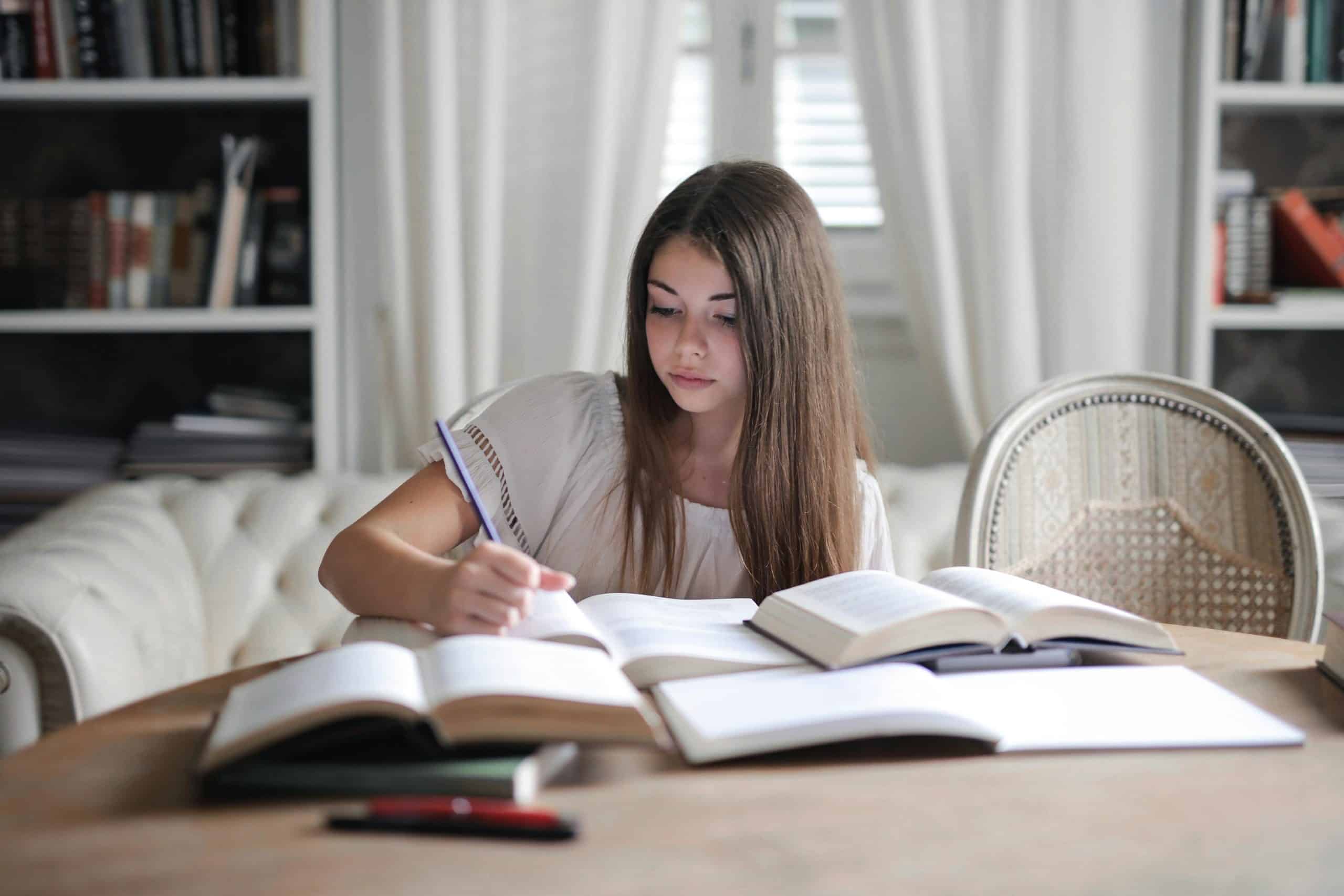 A high school student writes in a notebook while surrounded by open books, representing preparation for writing competitions for high school students.