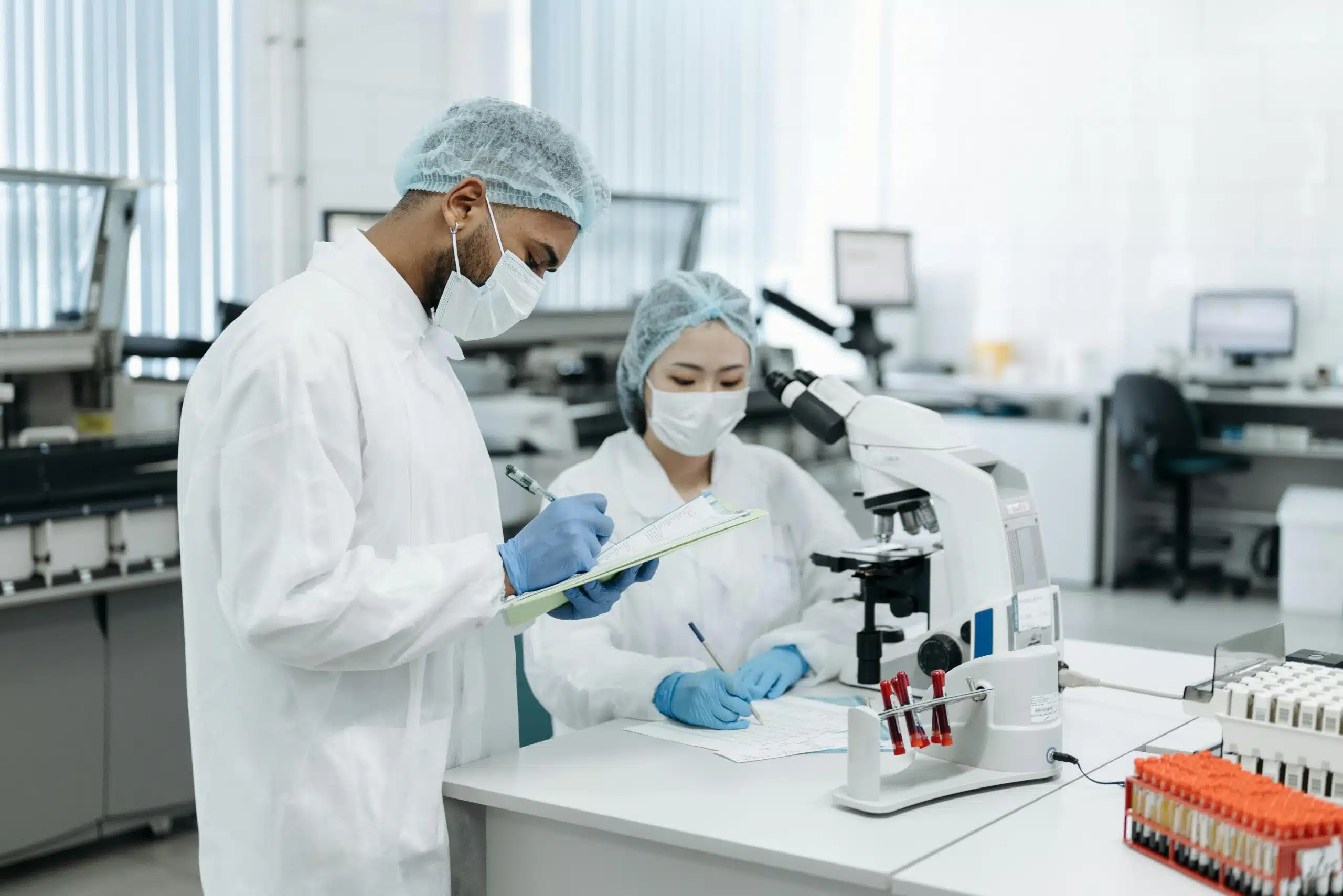 Two students in lab coats and masks working with a microscope and recording data, representing summer medical research programs for high school students.