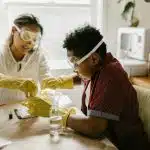 Adult and student wearing safety goggles and gloves conduct a hands-on experiment with liquids and lab tools at a tablerepresenting beginner-friendly chemistry research topics.