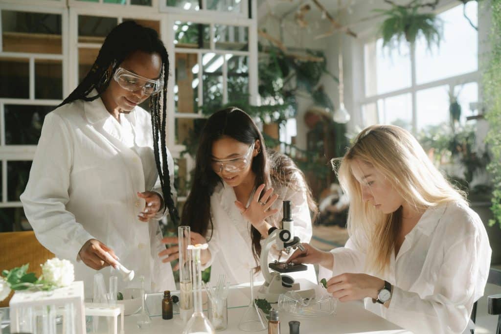 Three high school students work together on a lab experiment with microscopes and test tubes, representing science competitions for high school students.