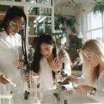 Three high school students work together on a lab experiment with microscopes and test tubes, representing science competitions for high school students.