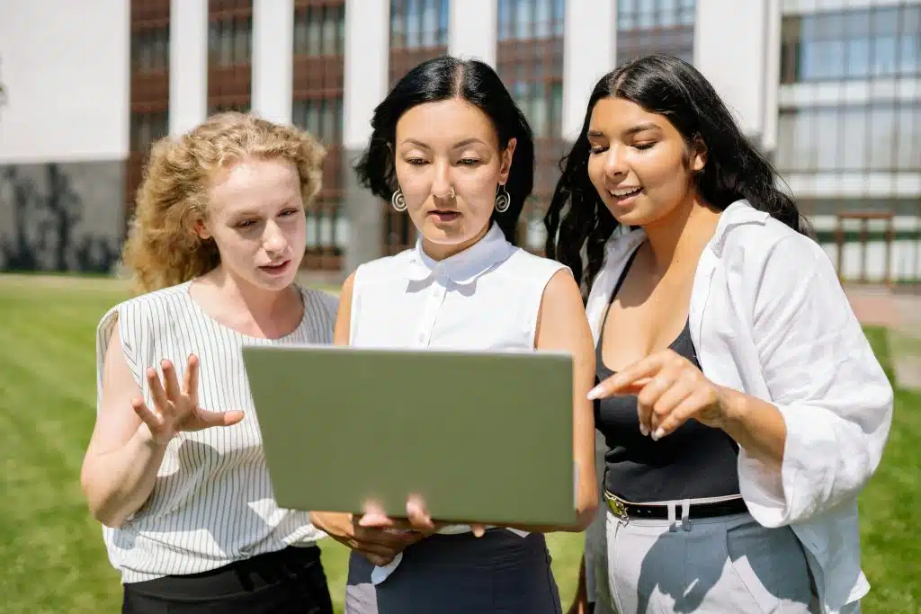 Three students standing outside on a college campus look closely at a laptop together, pointing at the screen as they discuss academic results and timelines, similar to students searching online for information like when do AP scores come out.