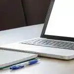 Open laptop with a blank screen next to a spiral notebook and two blue pens on a wooden desk — a Common App essay writing setup
