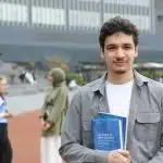 a student in a gray and white holding a blue book smiling after learning about the washu acceptance rate