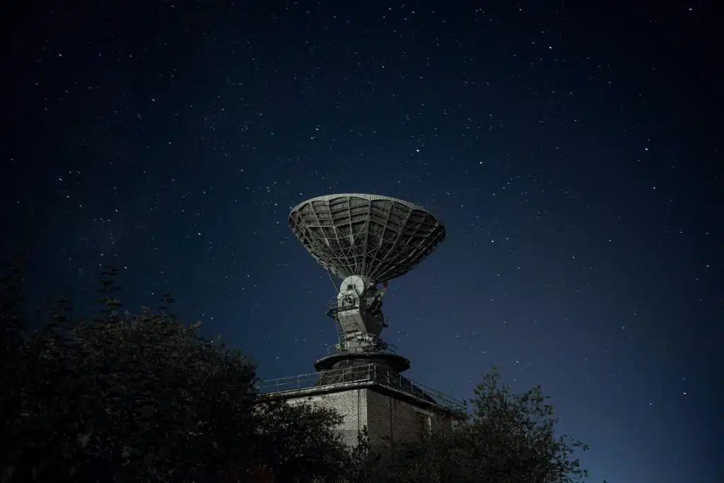 Large radio telescope dish against a star-filled night sky, surrounded by trees
