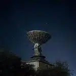 Large radio telescope dish against a star-filled night sky, surrounded by trees