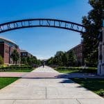 Purdue University entrance arch on a sunny day, with a tree-lined campus walkway and brick buildings beyond