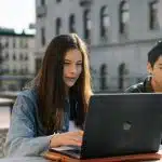 Two college students working on a laptop outdoors on campus, researching the University of Rochester acceptance rate