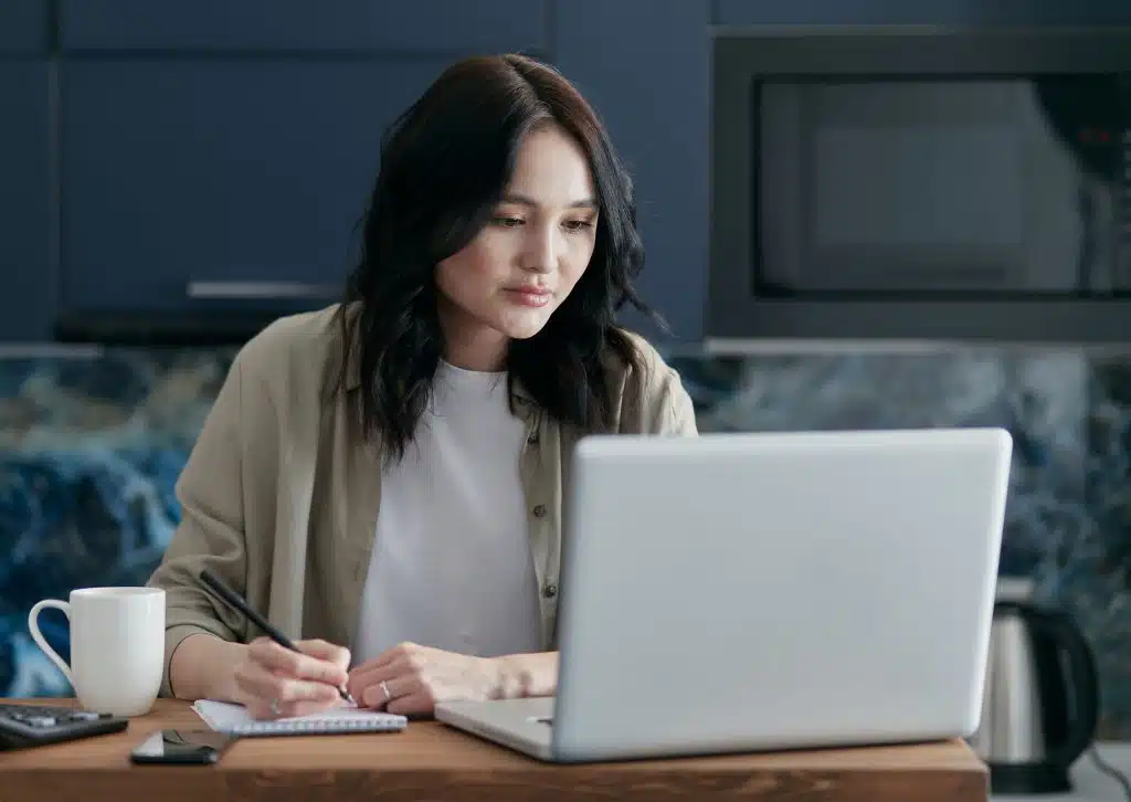 Person writing notes while working on a laptop at a kitchen table, researching and drafting a community essay with a coffee mug nearby.