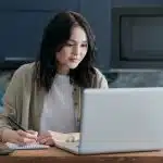 Person writing notes while working on a laptop at a kitchen table, researching and drafting a community essay with a coffee mug nearby.