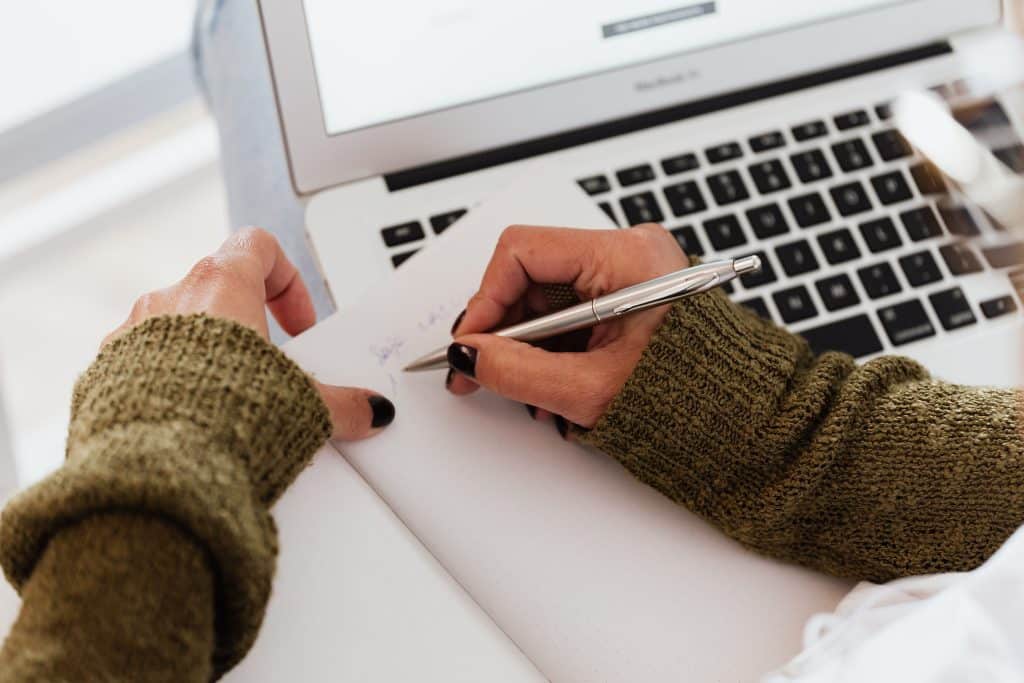 Student handwriting notes with a pen while planning QuestBridge essay prompts near a laptop