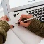 Student handwriting notes with a pen while planning QuestBridge essay prompts near a laptop