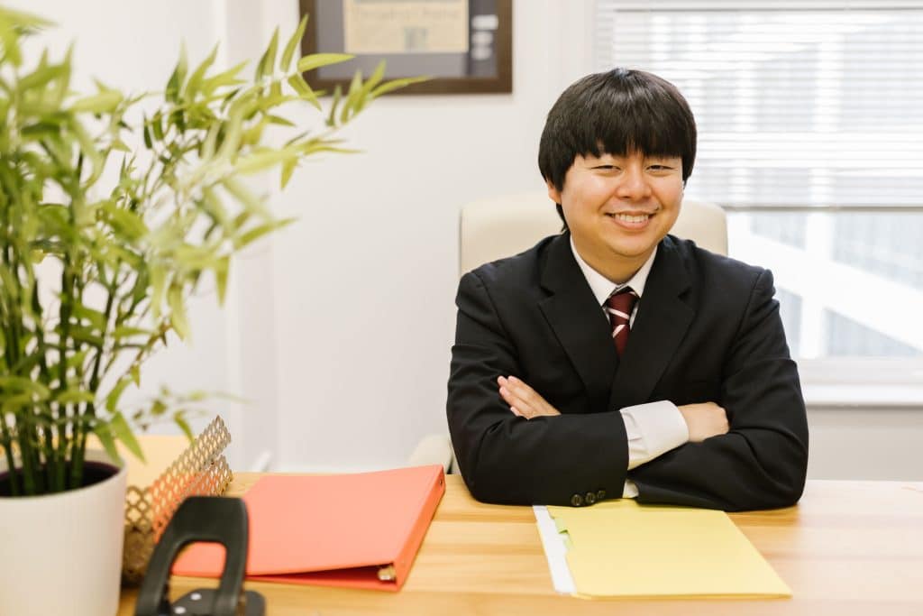 Yale admissions consultant in a dark suit smiling with arms crossed at a desk with a binder and folder